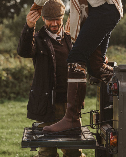 A person wearing tall brown riding boots and a brown flat cap stands on the tailgate of a vehicle outdoors while another person adjusts the hat. The scene is styled in a rustic, outdoor setting with grass and trees in the background.