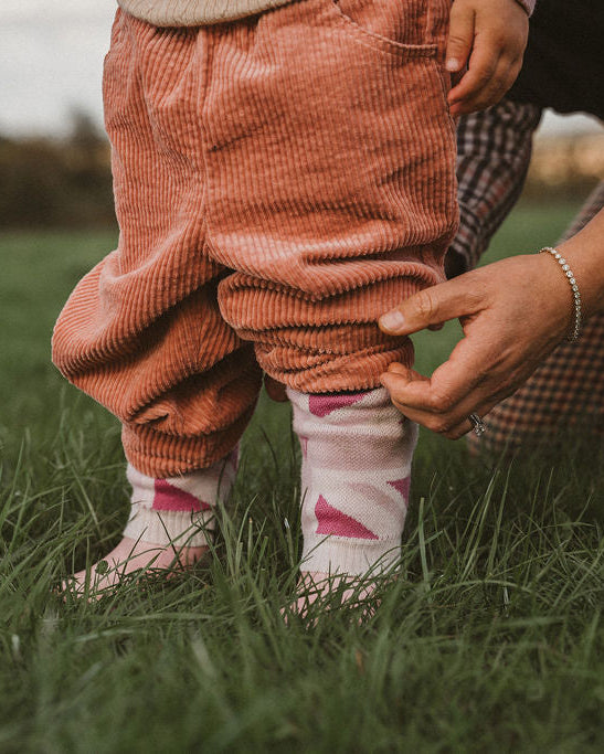 A close-up outdoor shot of a child wearing rust-colored corduroy pants and white socks with pink accents, while an adult in a plaid shirt adjusts the child's pants on a grassy field.