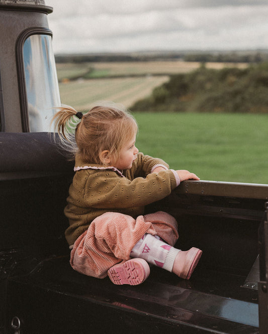 A young child sits in the back of a vintage black train, looking out over a green countryside landscape under a cloudy sky.