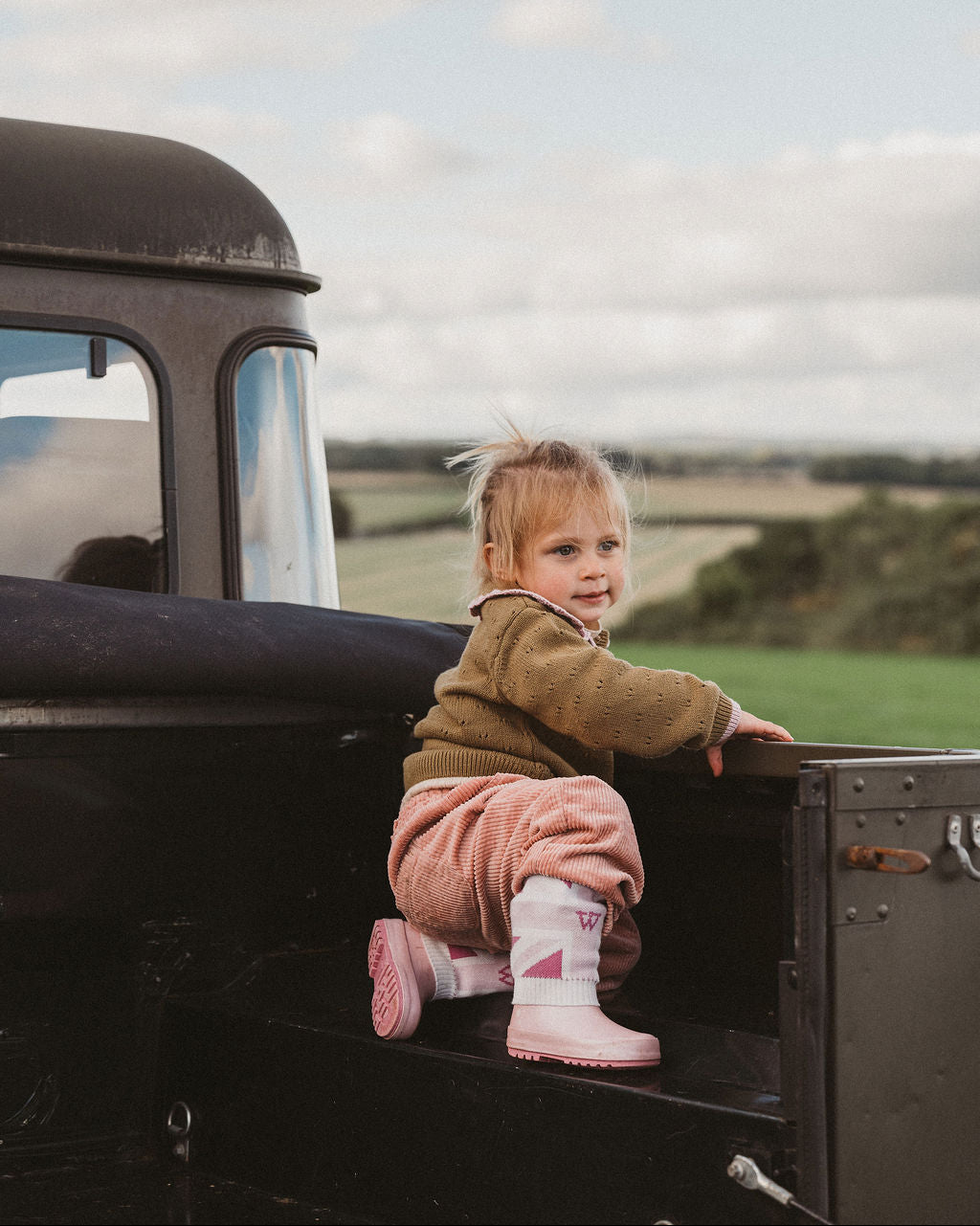 A toddler girl with blonde hair sits on the tailgate of a vintage black truck outdoors, wearing a brown knit sweater, pink pants, and pink boots. The background shows a soft-focus rural landscape under a cloudy sky.