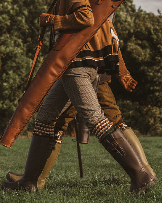 A person walking outdoors on grass wearing tall dark rubber boots, striped knee-high socks, and a brown jacket, while carrying a large brown leather bag or case.