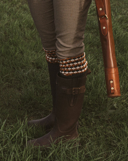 A close-up outdoor shot of a person's legs standing on green grass, wearing brown leather riding boots, patterned houndstooth trousers, and knit socks, with a brown leather gun stock or case held at the side.
