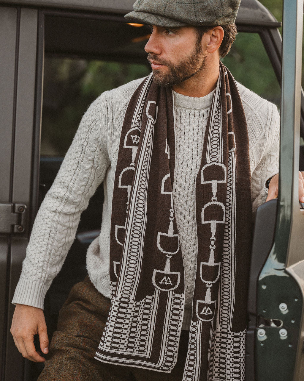 A man wearing a gray tweed flat cap, a light gray cable-knit sweater, brown tweed trousers, and a dark brown scarf with a white horse-bit pattern, leaning against a vintage green vehicle.
