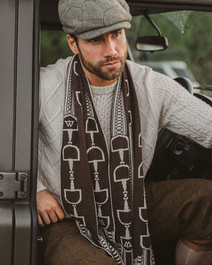 A man wearing a brown and white herringbone-patterned scarf, a gray flat cap, a light gray sweater, and brown tweed trousers, sitting inside a vehicle.